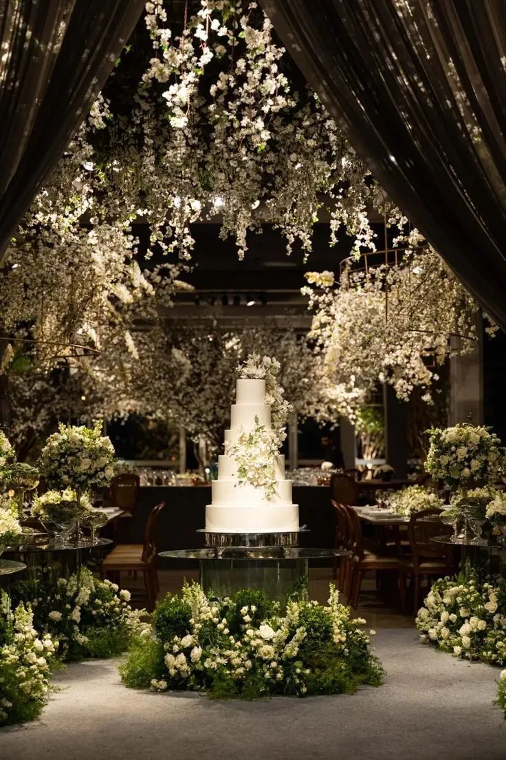 Decoração de casamento com bolos brancos e arranjos de flores brancas em ambiente elegante com teto decorado com flores.