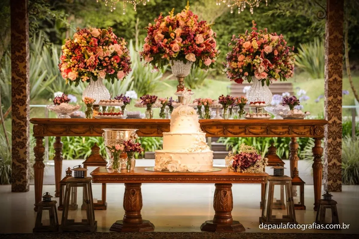 Mesa de festa de casamento decorada com flores e bolo em ambiente externo, ao fundo uma área verde. Decoração elegante e charmosa para celebração.