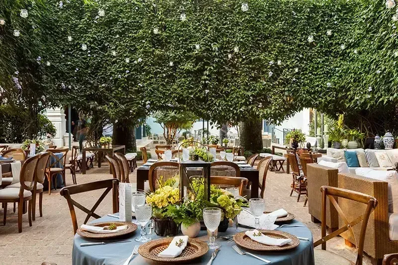 Imagem de um espaço ao ar livre decorado com mesas postas para um evento ou celebração, em um ambiente com árvores e decoração elegante.