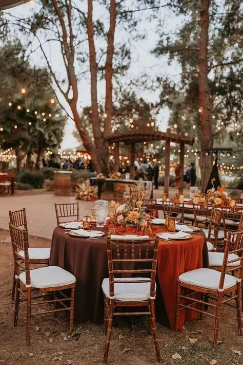 Mesa decorada ao ar livre com toalha marrom e detalhes sofisticados, ideal para eventos ao ar livre em ambiente natural com iluminação decorativa e árvores ao redor.