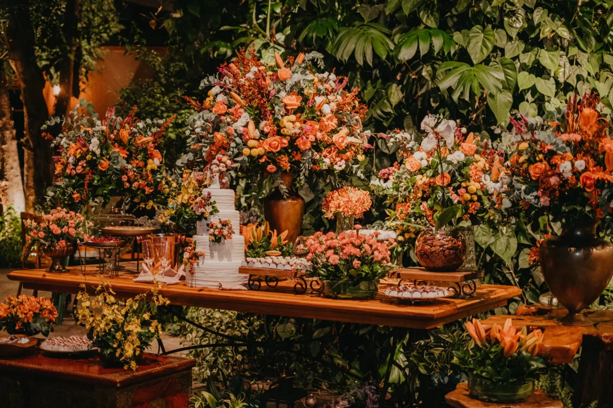 Decoração de festa com arranjos de flores em tons de rosa e laranja, mesas decoradas com doces e utensílios, ambiente com iluminação quente e cercado de plantas verdes.