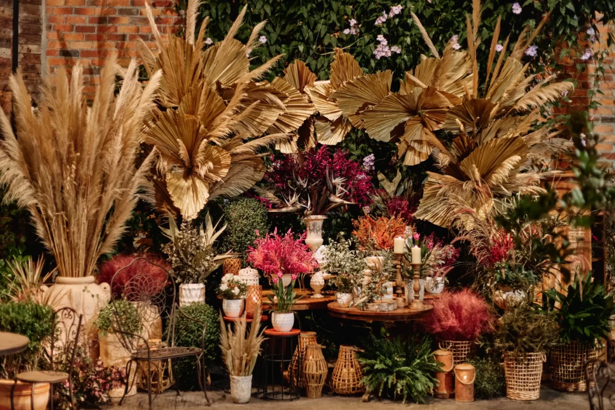 Arranjo de decoração com plantas e flores variadas, incluindo folhagens douradas, vasos de fibras naturais e velas, criando um ambiente rústico e elegante.