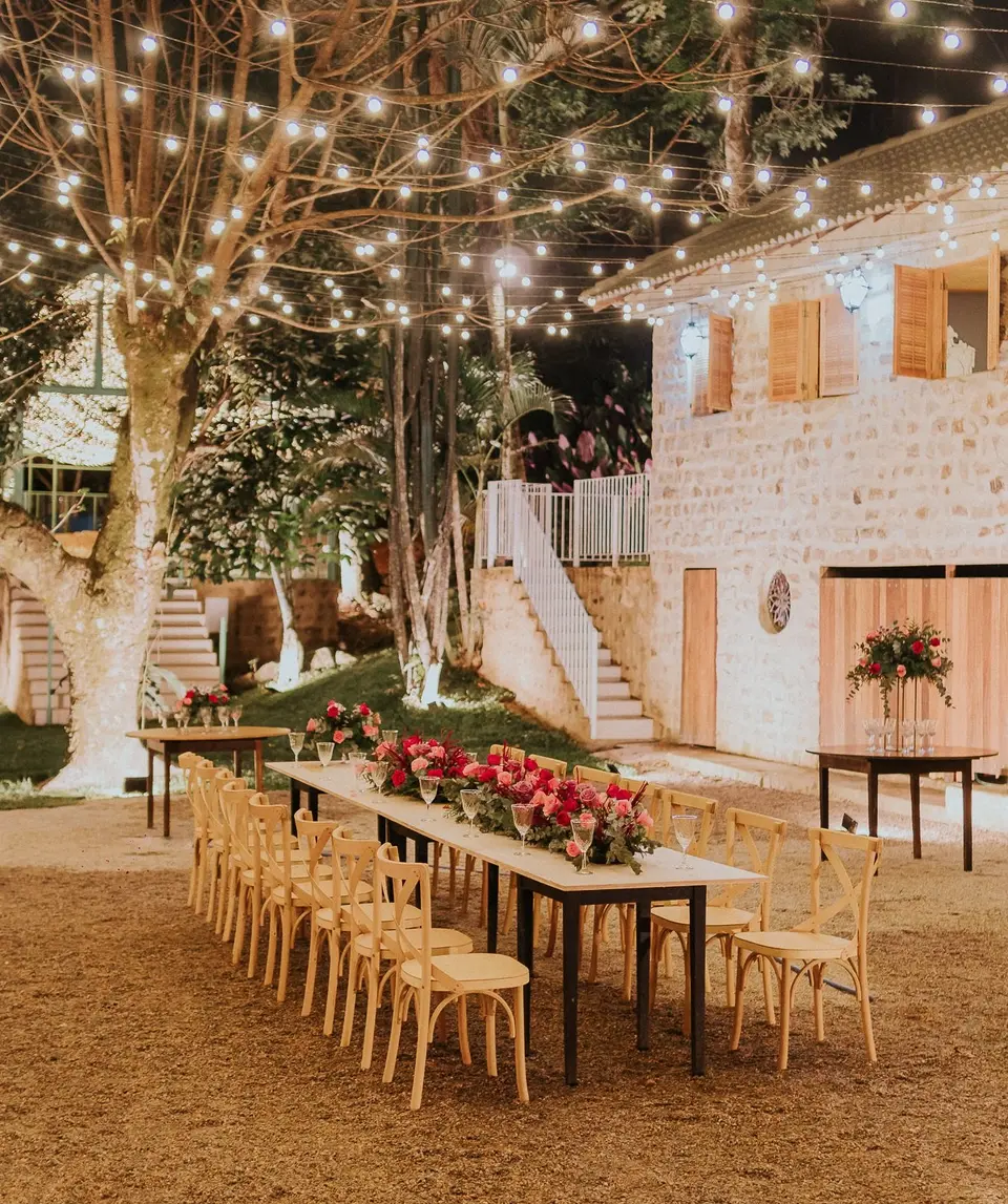 decoração de festa ao ar livre à noite com mesa de bolo e cadeiras decoradas, iluminada por luzes penduradas em árvores, cerimônia elegante em espaço ao ar livre