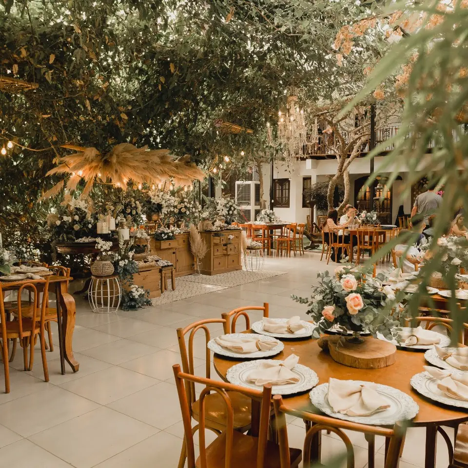 Imagem de uma festa ao ar livre decorada com flores e móveis de madeira, com destaque para mesas e cadeiras elegantes em um ambiente natural com árvores e iluminação suave.