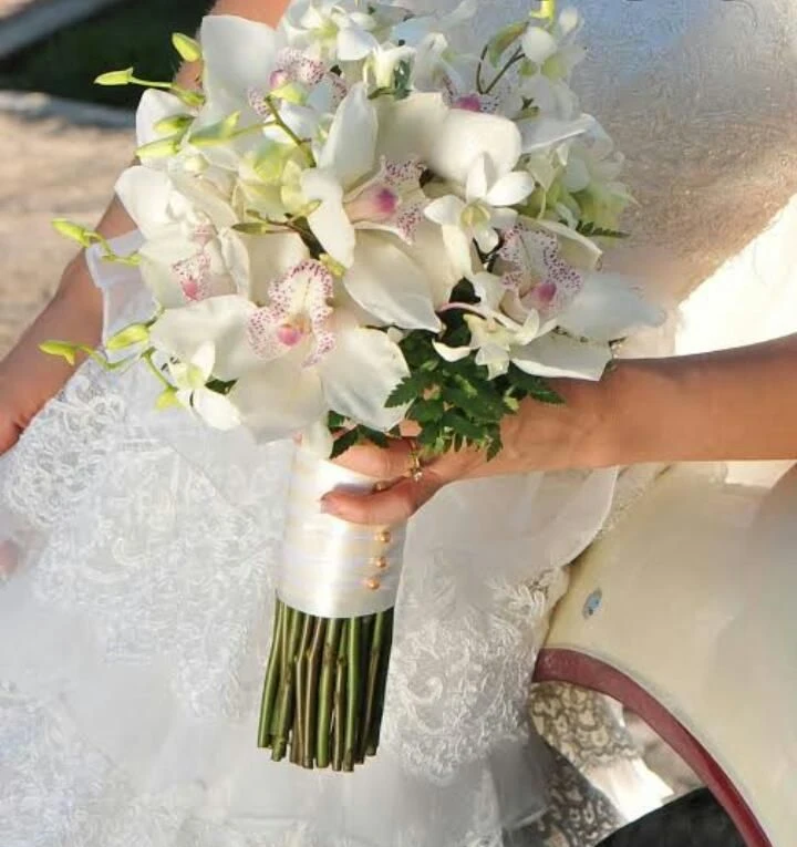 Buquê de flores brancas com detalhes em rosa, ideal para casamento, com mãos segurando na parte de baixo do vaso, ao lado de vestido de noiva.