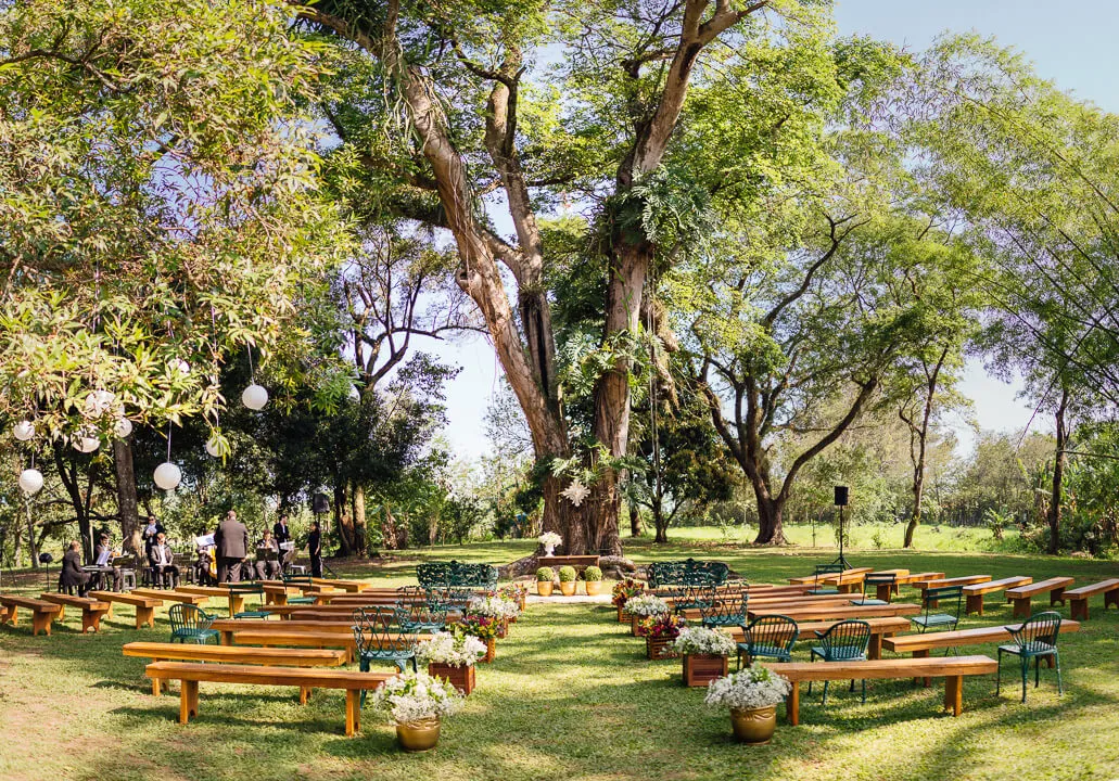 Cerimônia ao ar livre em parque com bancos de madeiras, cadeiras coloridas e muitas flores, sob grandes árvores verdes.