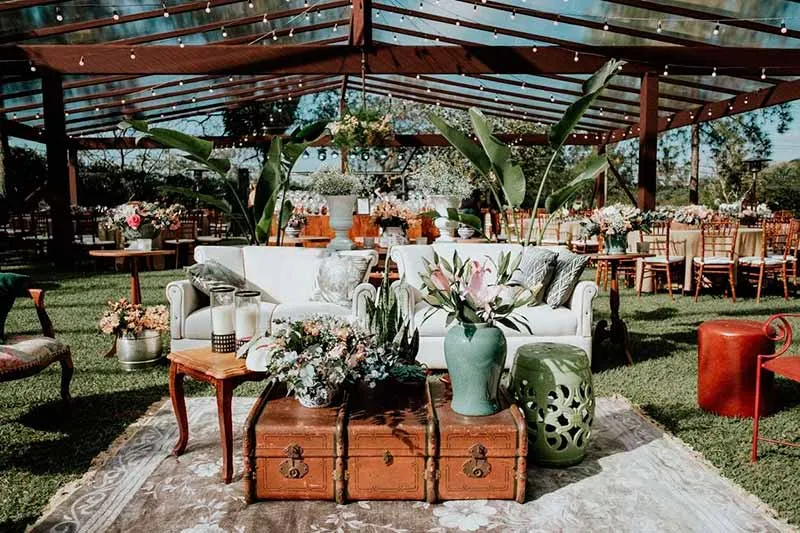 Decoração rústica com sofá branco, mesa de madeira, vasos com flores, toldo de madeira e iluminação de bistrô em evento ao ar livre.