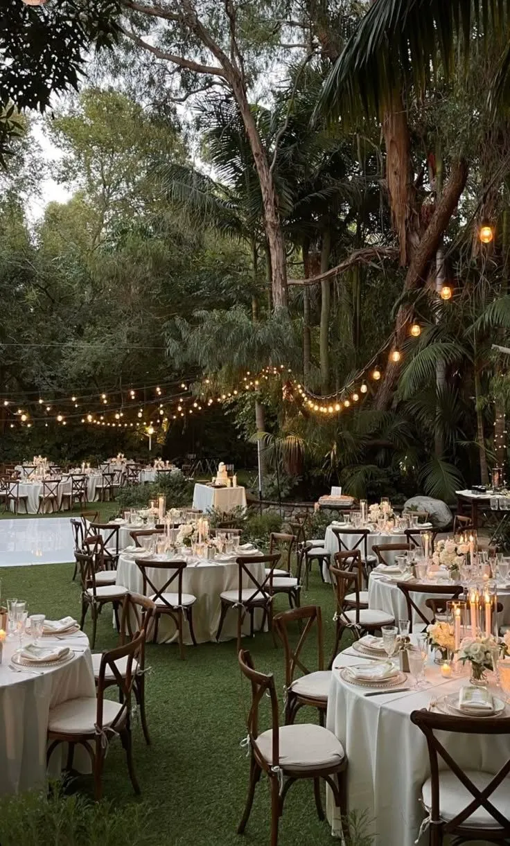 Imagem de um evento de casamento ao ar livre com mesas elegantes, cadeiras confortáveis, iluminação de lâmpadas de filamento e um ambiente rodeado por árvores e vegetação tropical.