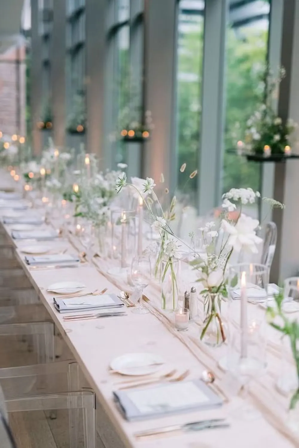 Decoração elegante de jantar com mesa decorada com flores brancas, velas e utensílios de prata em ambiente bem iluminado com janelas grandes.
