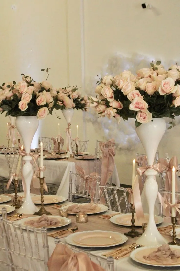 Decoração de mesa de casamento com arranjos sofisticados de rosas em vasos altos brancos, velas acesas e detalhes em cetim em tom nude, ideal para eventos elegantes.