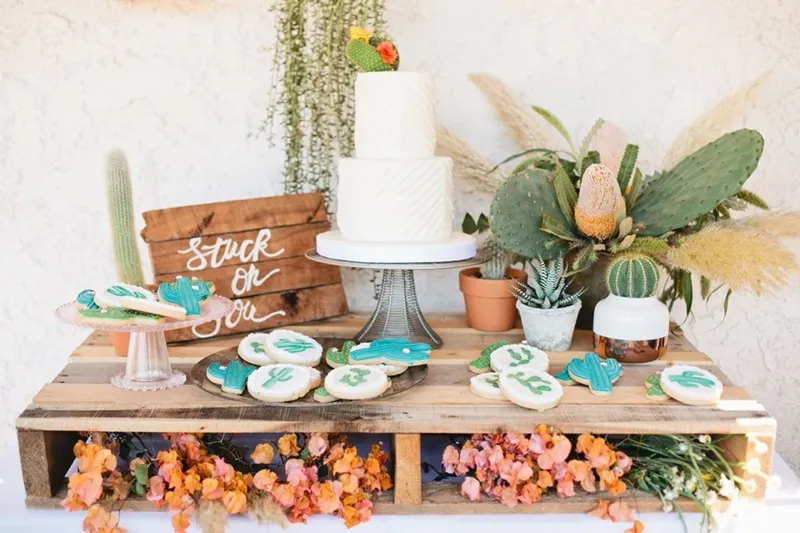 Decoração de festa com bolo branco, cookies decorados, arranjos de flores e plantas suculentas em um ambiente rústico e elegante.