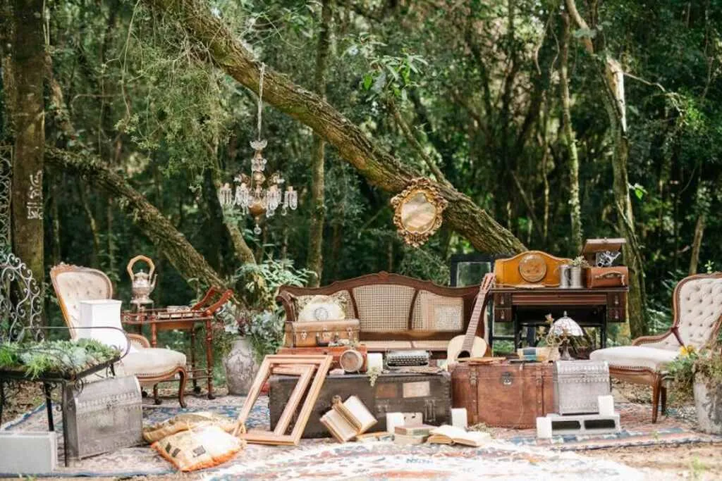 Decoração vintage ao ar livre em meio à natureza, com móveis antigos, instrumentos musicais e itens decorativos em um cenário de floresta.