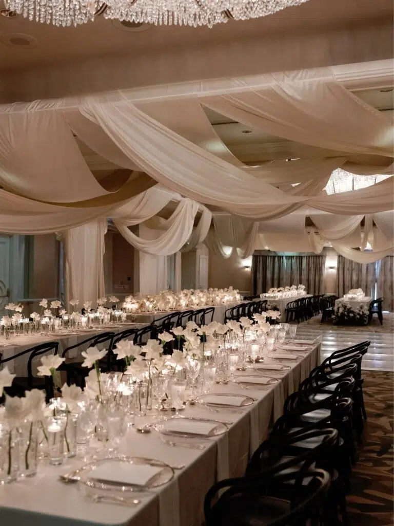 Decoração elegante para evento sofisticado com teto de tecido drapeado, longa mesa de jantar com arranjos de flores brancas e velas em ambiente bem iluminado