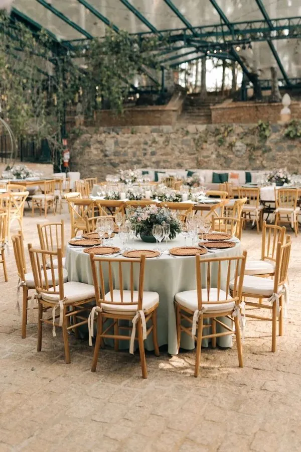 Imagem de mesas de jantar bem decoradas em um espaço ao ar livre para eventos ou festas, com cadeiras de madeira e centro de mesa com flores brancas em um ambiente com teto de vidro e paredes de pedra.
