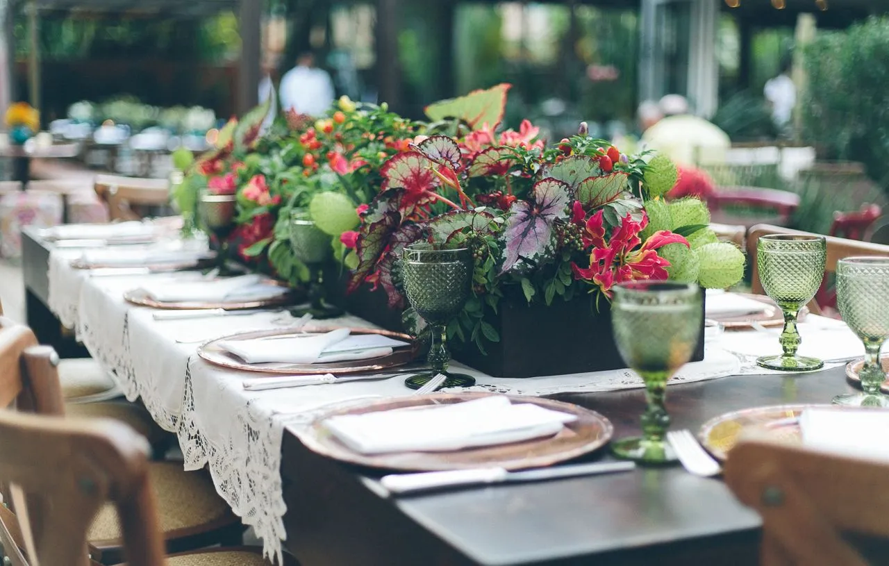 Mesa de jantar ao ar livre decorada com flores e taças de vidro, preparada para um evento ou celebração ao ar livre em um ambiente natural e elegante.