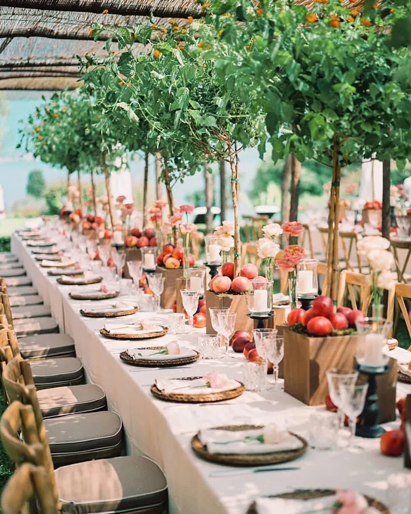 Decoração de festa ao ar livre com mesa grande, árvores decorativas, frutas, flores e copos elegantes, ideal para casamento ou evento familiar