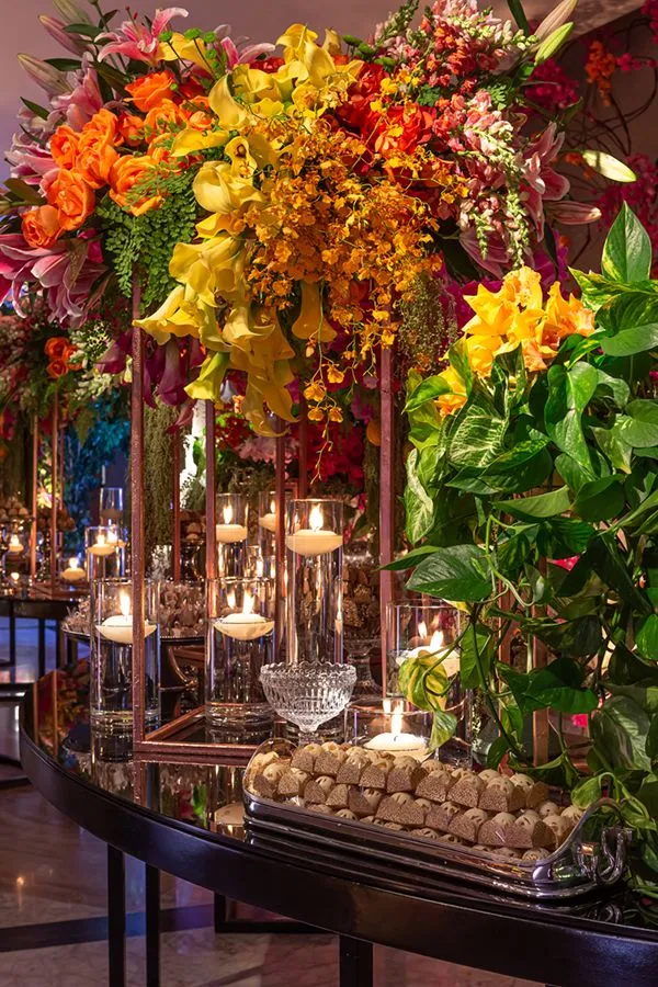 Arranjo de flores vibrantes com rosas, orquídeas e folhagens, decorando uma mesa de evento elegante com velas acesas e elementos de cristal.