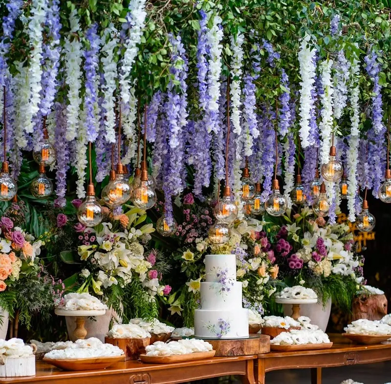 Decoração de casamento com flores penduradas em tons de lilás e branco, iluminação de lâmpadas expostas e bolo decorado com detalhes florais.