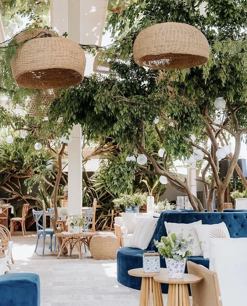 Interior de um espaço aconchegante com decoração natural, incluindo árvores, luminárias de teto de fibra natural, poltronas azuis e mesas com flores, criando um ambiente relaxante e elegante.