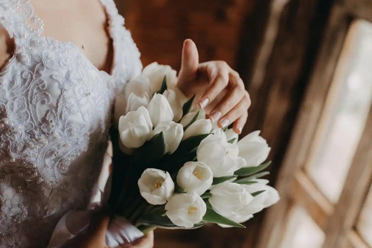 Mulher em vestido de casamento segurando um buquê de tulipas brancas ao lado de uma janela, simbolizando um momento especial de casamento.