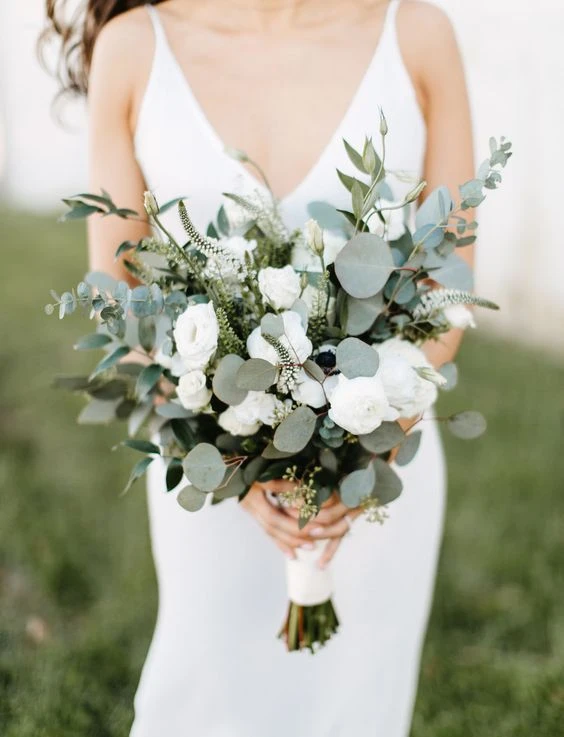 Mulher com vestido branco segurando buquê de flores naturais, composição com folhas verdes e flores brancas, cenário ao ar livre.