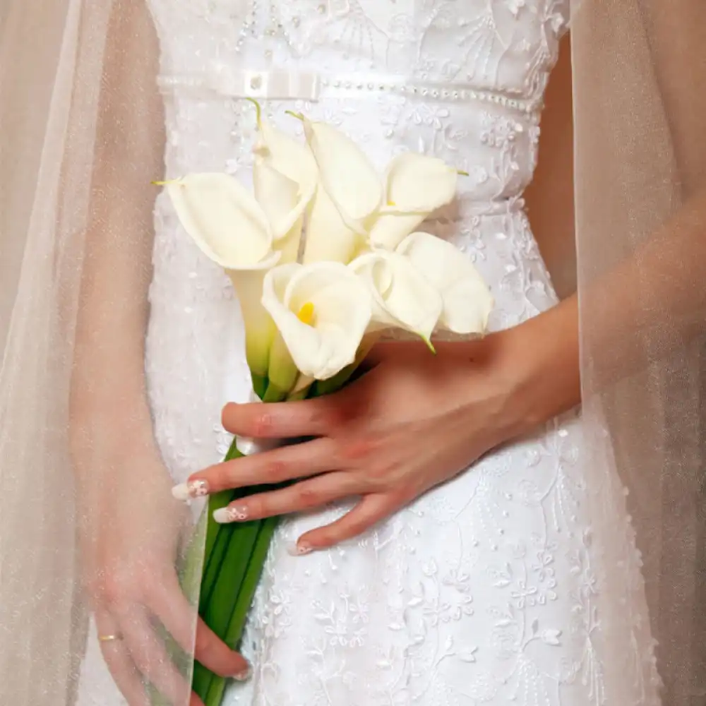 Imagem de uma noiva segurando um buquê de calas brancas em um vestido de casamento com detalhes em renda bordada, símbolo de noivado ou casamento.