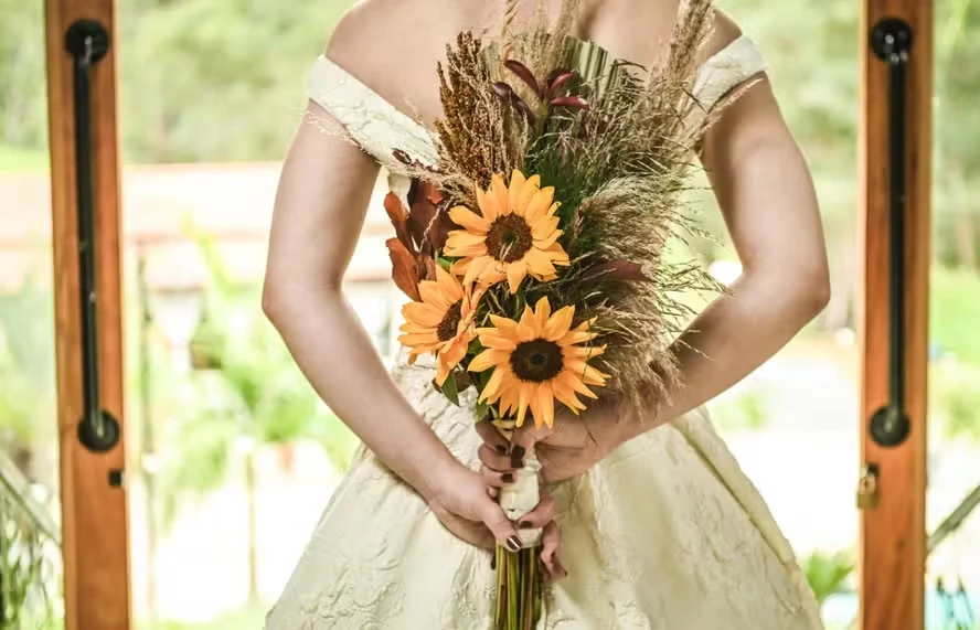 Imagem de uma noiva segurando um buquê de flores composto por girassóis, ao fundo uma porta de madeira com detalhes decorativos e ambiente verde ao ar livre
