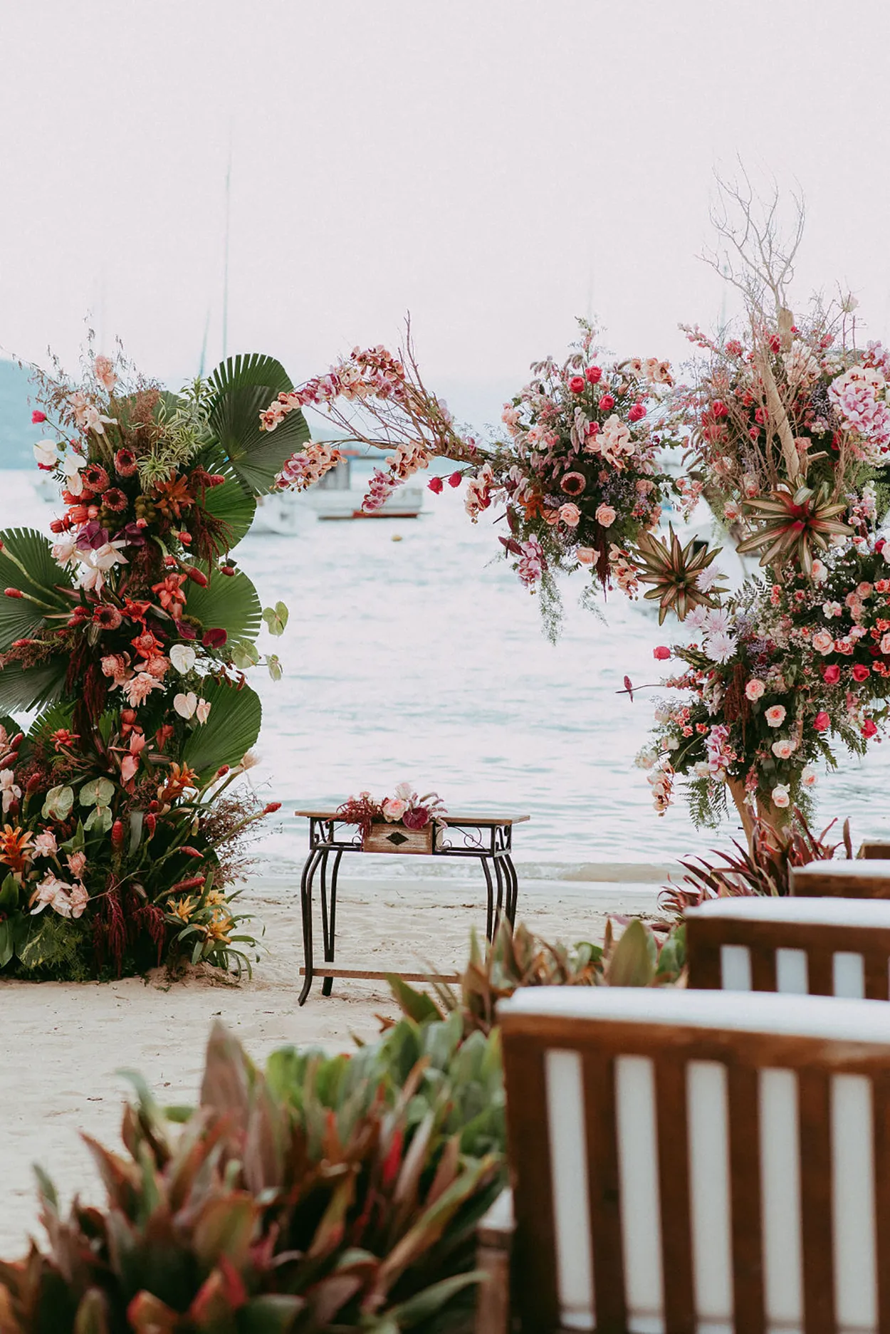 Decoração de casamento na praia com arranjos florais rosa e verde, vista para o mar ao fundo, ideal para celebrações ao ar livre.