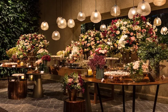Decoração elegante com várias arranjos de flores coloridas em tons de rosa, branco e roxo, iluminados por luminárias de teto e velas, ideal para eventos sofisticados.