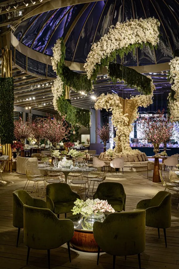 Decoração luxuosa para evento com detalhes em flores brancas e rosa, iluminação elegante e ambiente sofisticado com cadeiras confortáveis e janelas amplas.