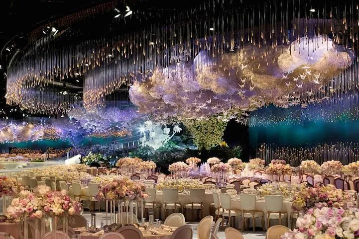 Decoração elegante para casamento com teto decorado com iluminação e nuvens de luz, mesas decoradas com arranjos de flores em ambiente sofisticado.