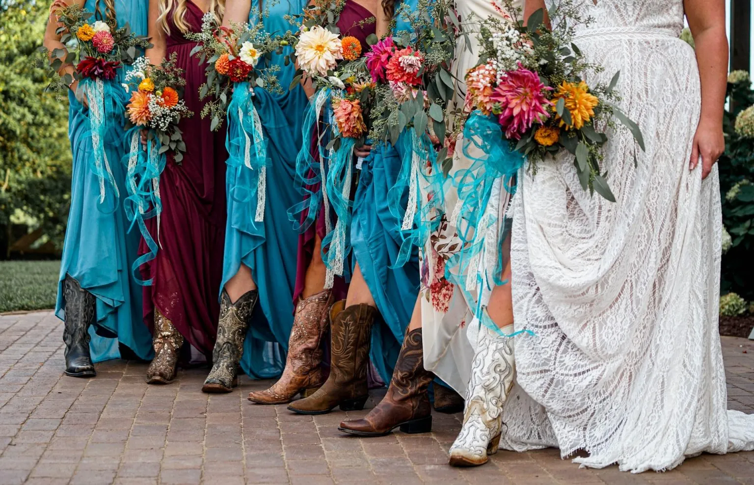 Mulheres vestidas de vestidos longos com botas cowboy, segurando buquês de flores, preparando-se para a cerimônia de casamento ao ar livre.
