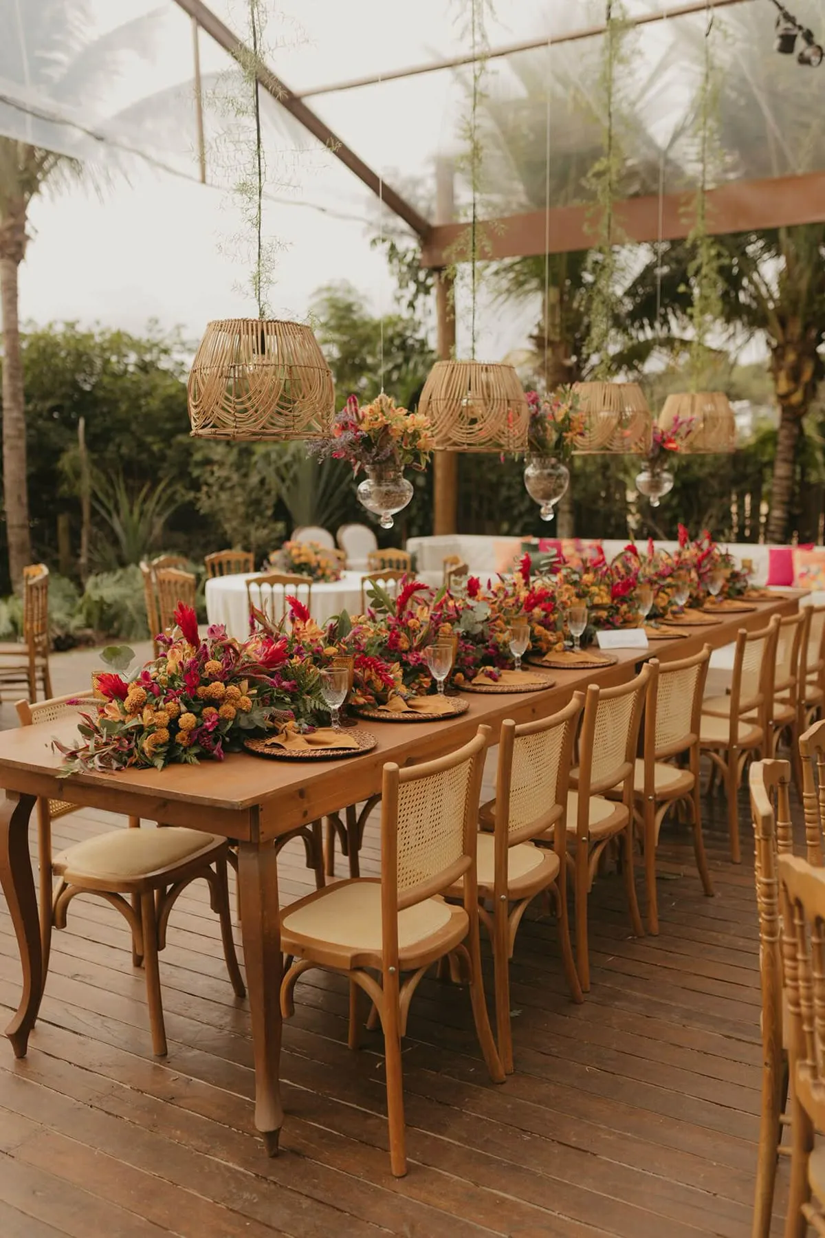 Imagem de uma mesa de jantar decorada com flores vibrantes, com luminárias de bambu suspensas, ambiente externo elegante ideal para eventos ao ar livre.