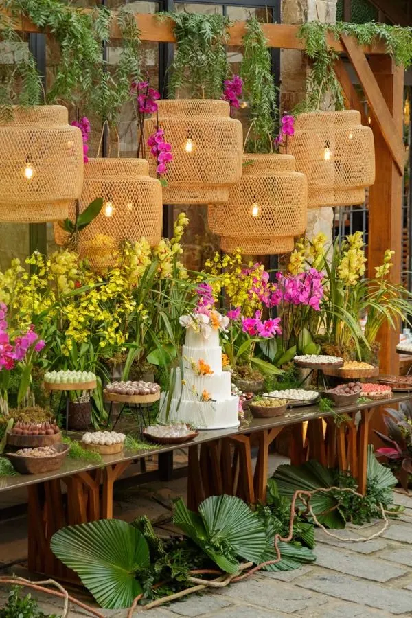 Decoração de festa com mesas cheias de doces, bolo branco decorado com flores, e arranjos de flores vivas, incluindo orquídeas, rodeada de plantas verdes e iluminação aconchegante em ambiente ao ar livre.