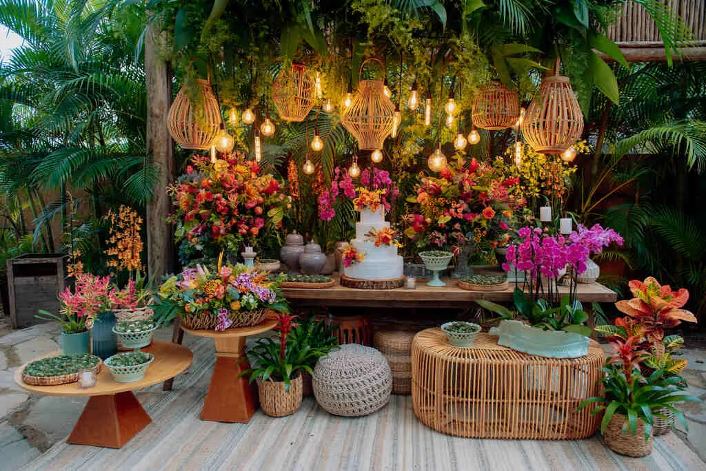 Decoração de festa ao ar livre com flores vibrantes, luzes penduradas, bolo de casamento, móveis de vime, ideal para celebrações especiais ao ar livre.