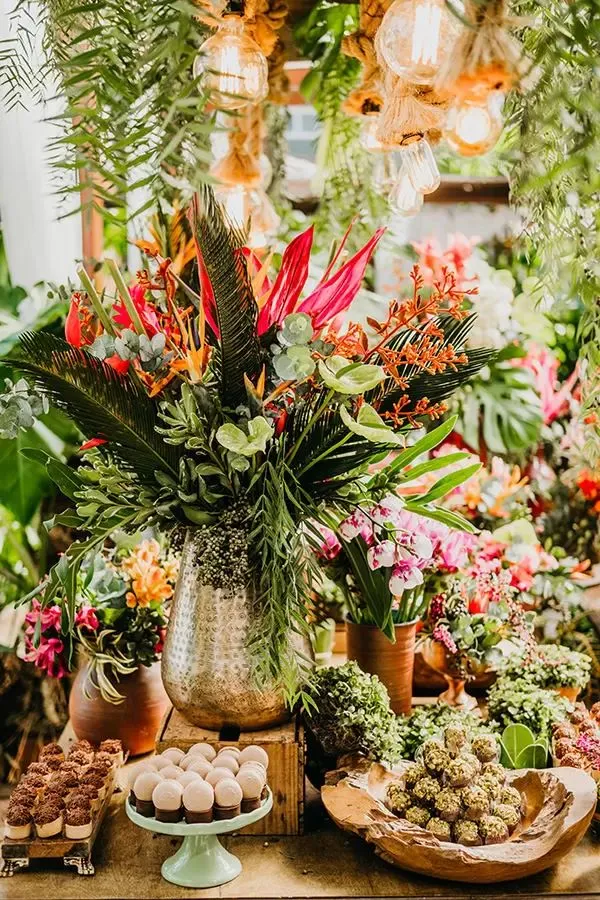 Arranjo de flores com diversas plantas e flores coloridas, incluindo folhas verdes e galhos em um ambiente bem decorado para celebrações ou eventos.