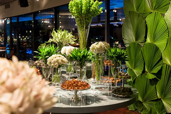 Decoração elegante com arranjos de flores brancas em vasos de vidro na mesa de evento ao lado de paredes cobertas por folhagens verdes, ambiente sofisticado e bem iluminado.