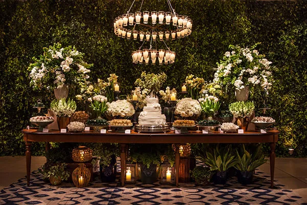 Decoração de casamento com um bolo central, flores brancas, velas, iluminação suave e arranjos exuberantes em um ambiente elegante ao ar livre, ideal para festas especiais.