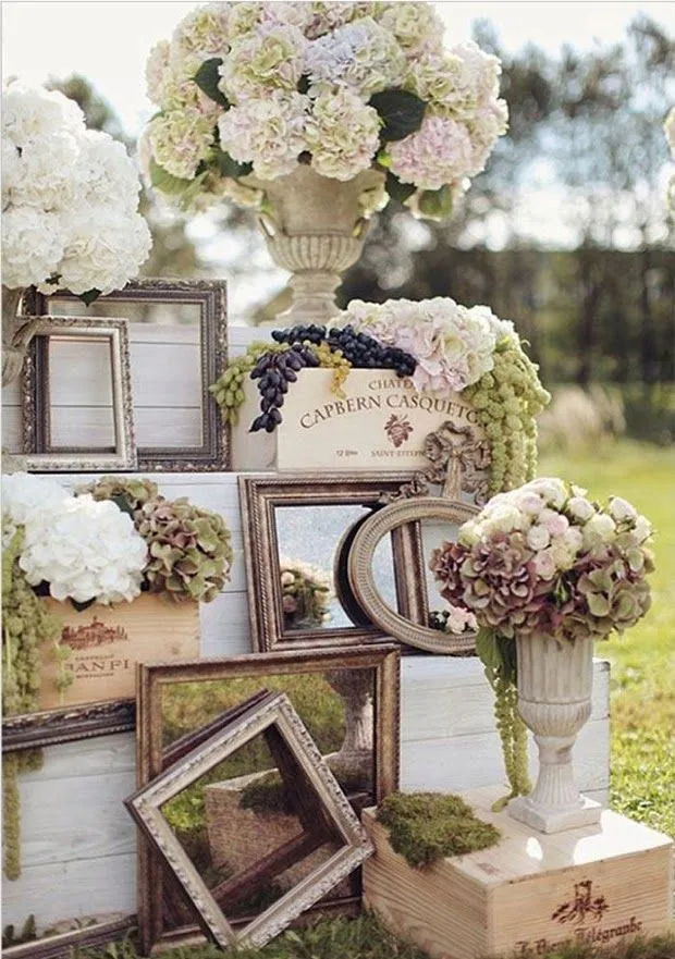 Arranjo de flores brancas, lilás e verdes em vasos clássicos, com molduras de quadros decorativos vintage, numa decoração de casamento ao ar livre.