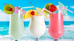 Três coquetéis coloridos com frutas decorativas em uma praia com céu azul ao fundo, ideal para consumo em dias quentes e festas à beira-mar.