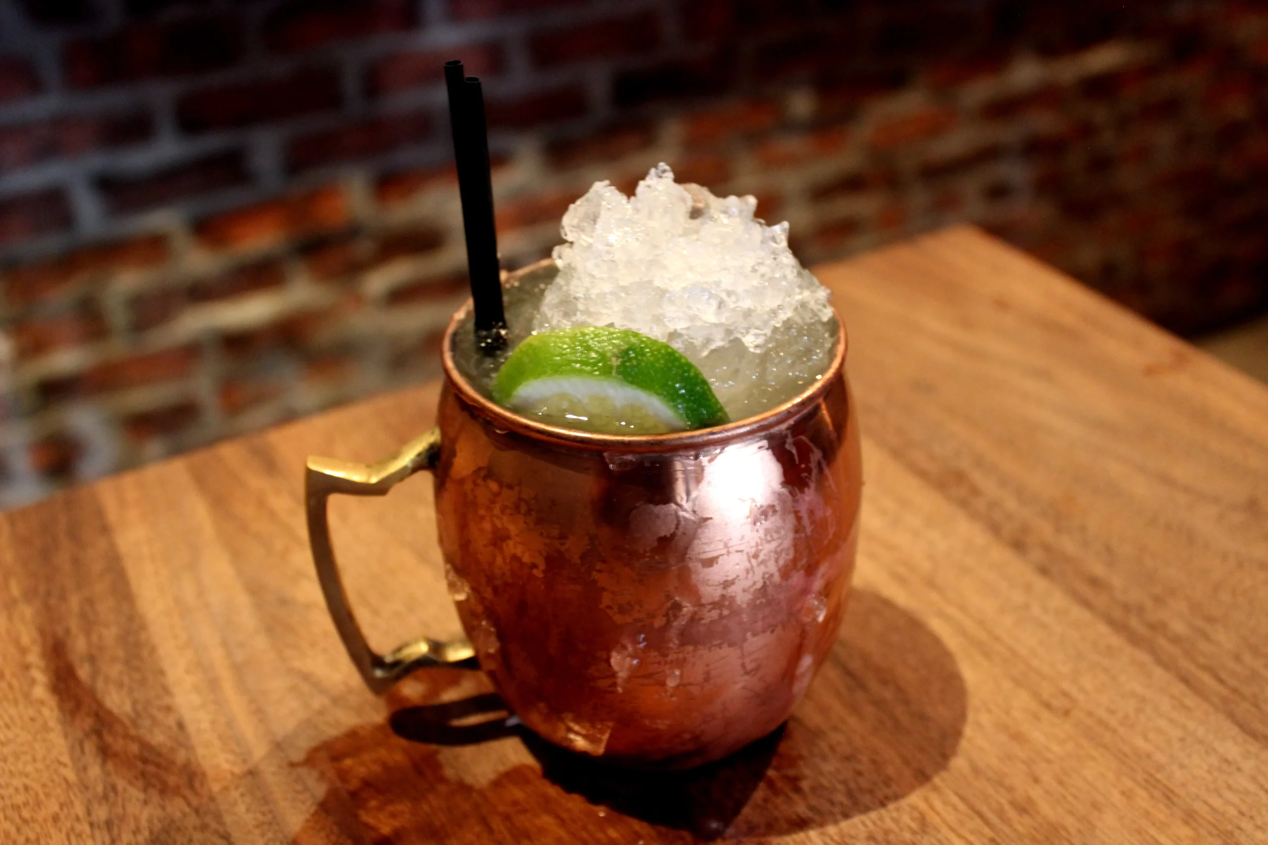 Coquetel de caipirinha com limão, gelo e canudo em copo de metal, servido em mesa de madeira com fundo de parede de tijolos.