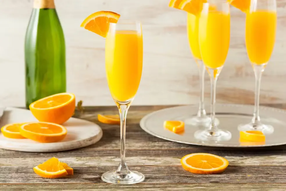Copo de espumante com suco de laranja decorado com fatia de laranja, ao lado de garrafa de espumante e fatias de laranja, ideal para festas e celebrações.