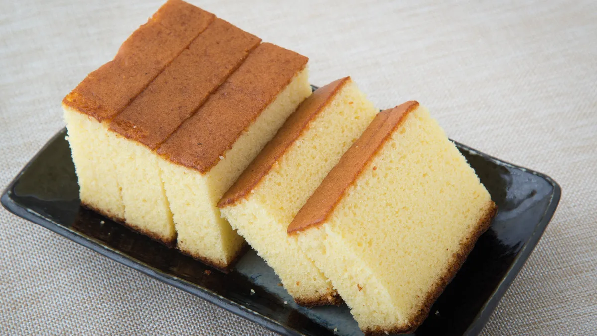 Três fatias de bolo de caixinha com cobertura dourada, dispostas em um prato preto sobre uma mesa cinza. O bolo tem aparência macia e esponjosa, ideal para lanches.