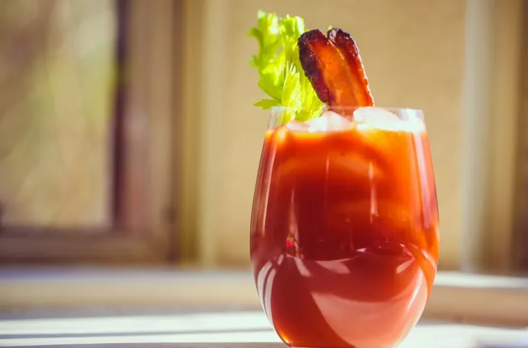 Imagem de um coquetel Bloody Mary decorado com talos de alface, uma fatia de bacon e um pedaço de limão, servido em um copo transparente.