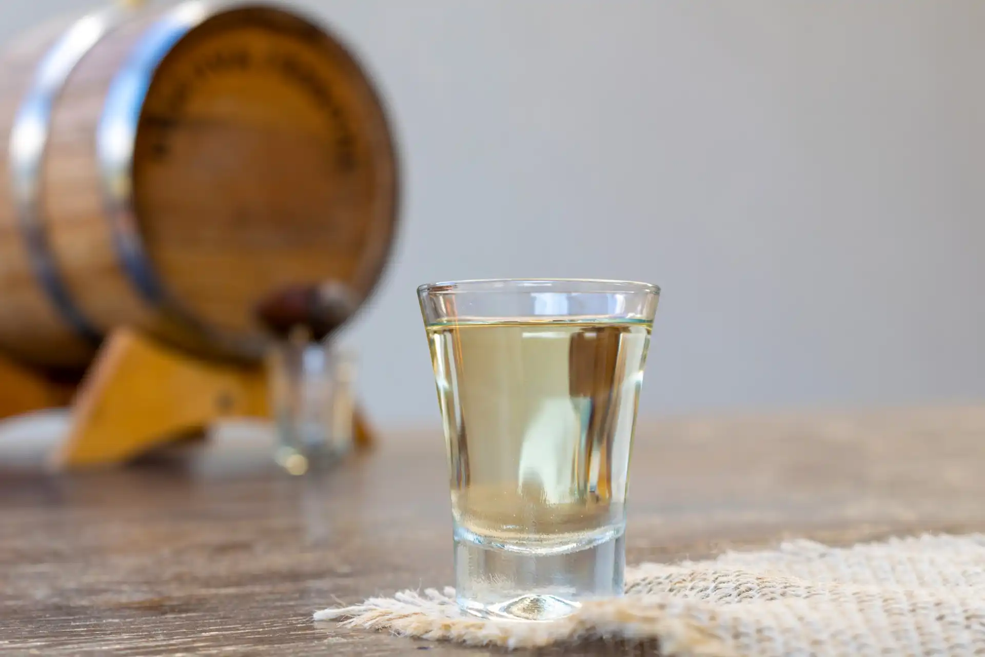 Copo de cachaça transparente em mesa de madeira com barril ao fundo, símbolo típico da bebida popular brasileira.