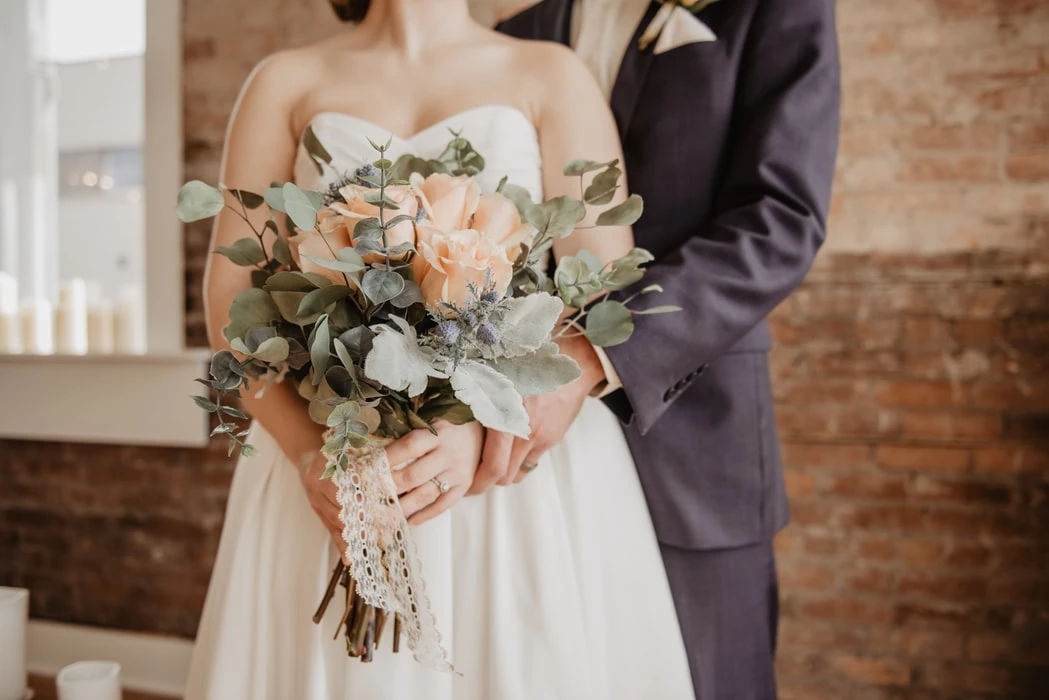 Casal de noivos segurando buquê de flores romântico, em cerimônia de casamento, com decoração rústica de parede de tijolos ao fundo.