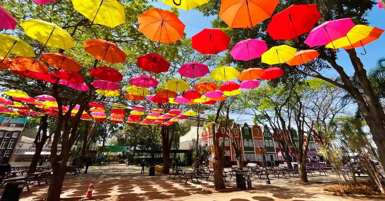 Decoração de parque urbano com guarda-chuvas coloridos suspendidos entre árvores, criando um ambiente vibrante e alegre ideal para fotos e eventos ao ar livre