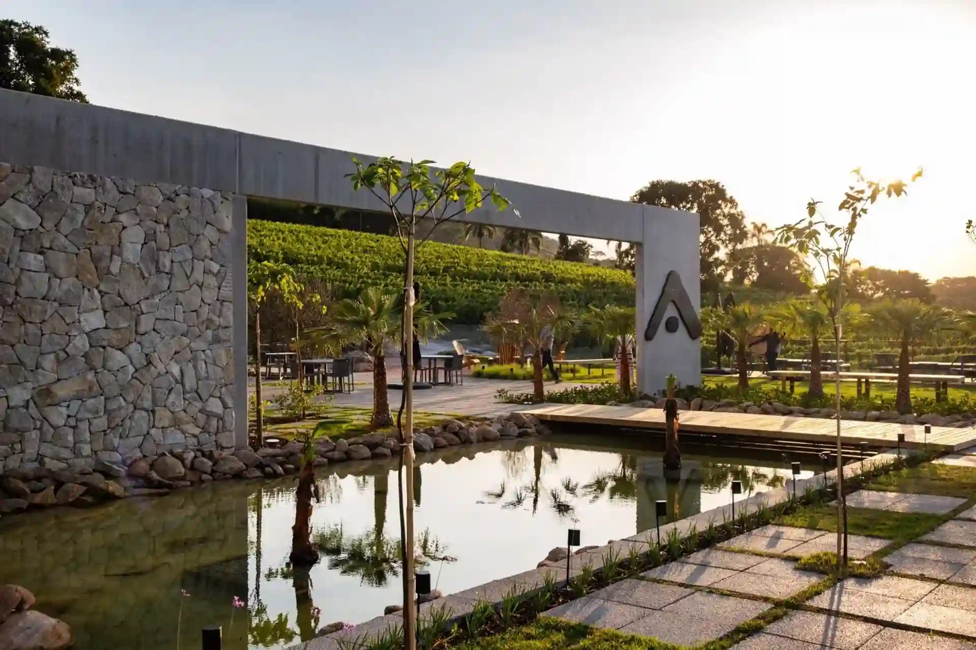 Imagem de um espaço ao ar livre com um lago refletindo árvores, um arco de entrada moderno e um cenário de paisagem com vegetação, ideal para área de lazer ou eventos.