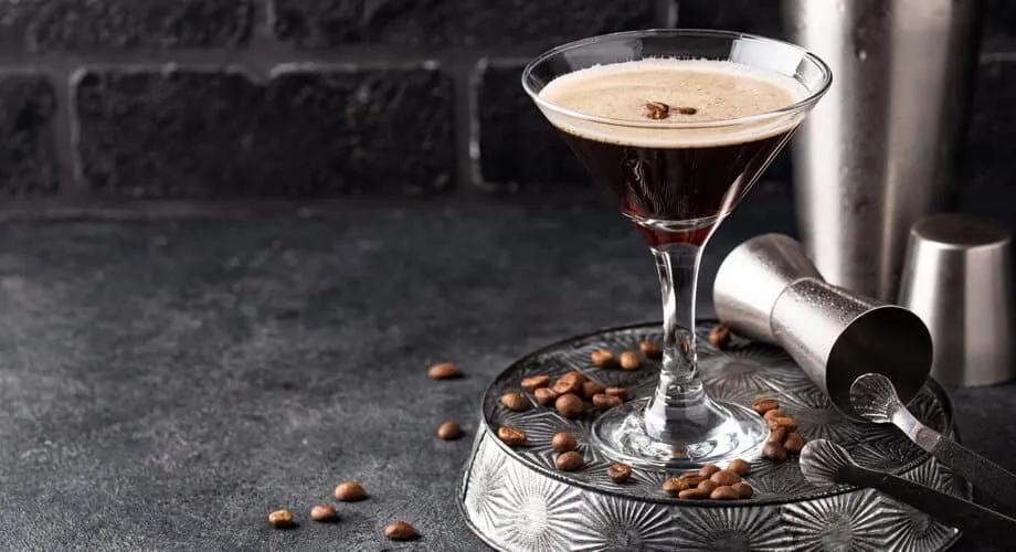 Copo de coquetel espresso martini em um ambiente sofisticado, decorado com grãos de café ao redor, com utensílios de bar ao fundo.