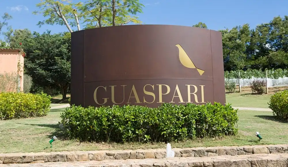 Entrada do empreendimento Guaspari com logo de passarinho dourado e nome em destaque, cercada por vegetação verde e jardins bem cuidados.